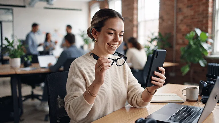 Lächelnde Frau sitzt im Büro und hält eine Meta AI Brille sowie ein Smartphone in der Hand auf dem sie die neuen Instagram Story Formate anschaut