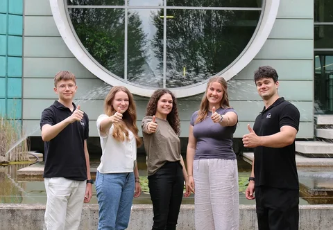 Fünf Auszubildende der Krick Unternehmensfamilie stehen vor dem Firmengebäude, halten den Daumen hoch und lachen in die Kamera