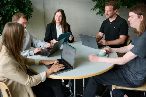 Fünf Personen sitzen mit Laptops um einen Tisch herum, unterhalten sich und lächeln