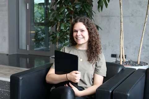 Junge Frau sitzt in einem Sessel mit Laptop in der Hand und lächelt in die Kamera