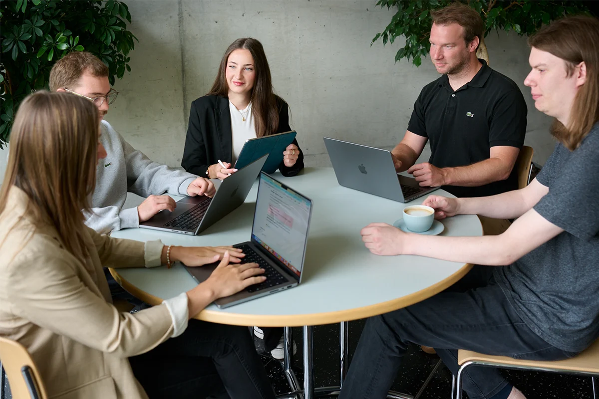 Fünf Personen sitzen mit Laptops um einen Tisch herum, unterhalten sich und lächeln